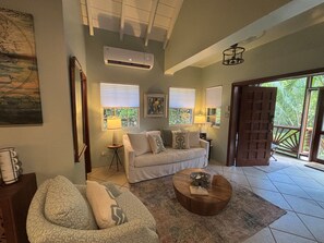A living room with a sofa, a coffee table, and a door leading to a balcony. - Sol Cottage Villa 2 BR 1 BA private pool lush landscaping ACs & new generator (Cruz Bay)