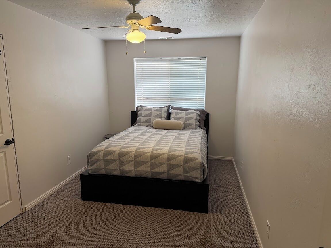 A bedroom with a bed, a ceiling fan, a window with blinds, and a door.