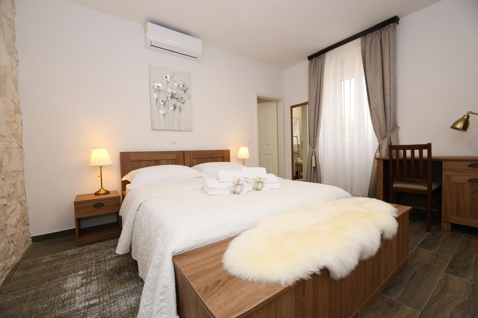 Un dormitorio con cama de madera, dos mesitas de noche con lámparas, una silla, un escritorio, aire acondicionado y un espejo.