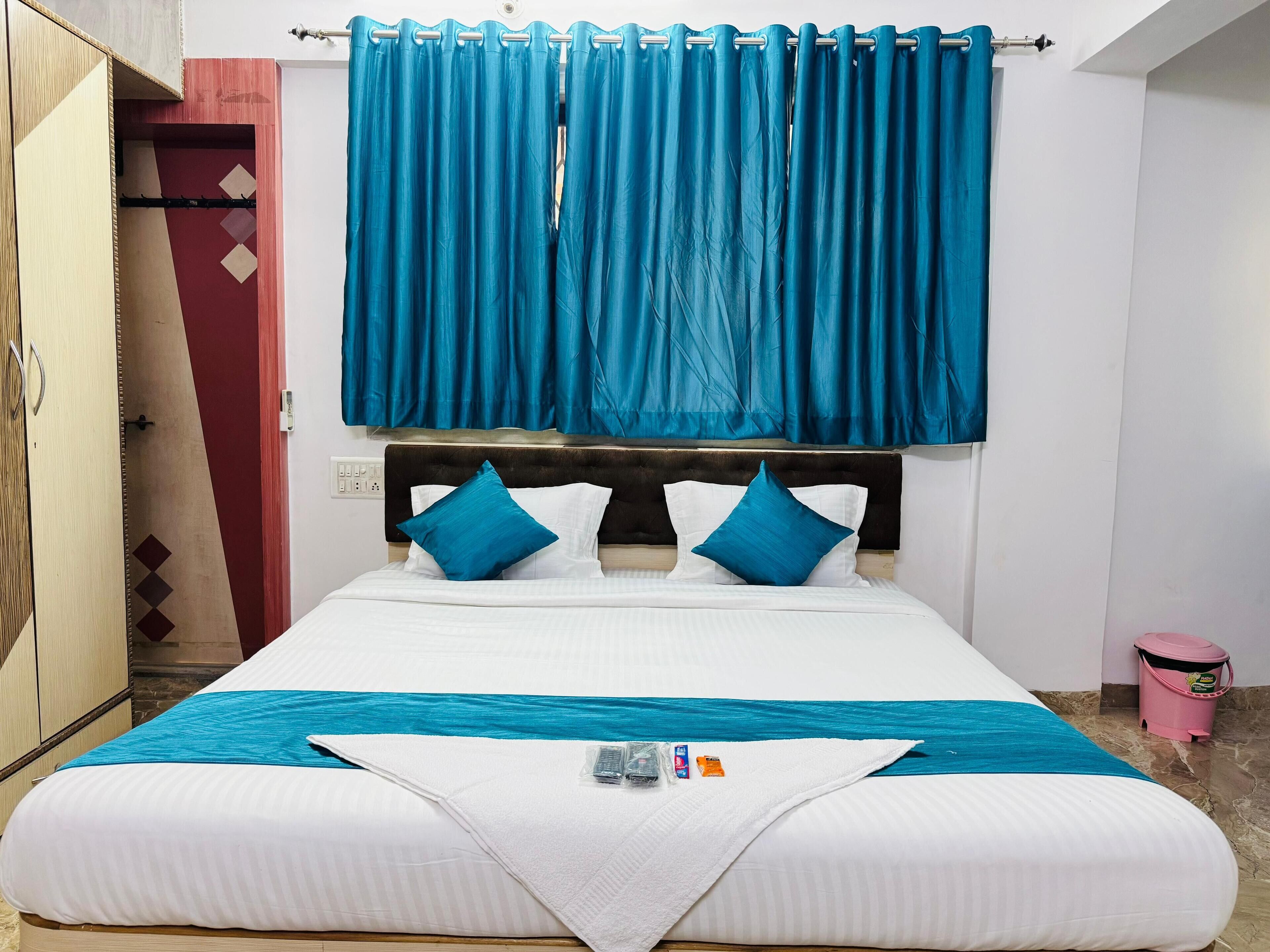 A neatly made bed with a blue and white bedspread, two blue pillows, and a white pillow. A blue curtain is drawn over the window, and a pink bucket is visible on the floor.