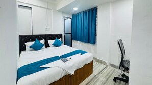 A neatly made bed with a white and blue bedspread, a wooden headboard, and a blue curtain on a window. - Hotel Raksha Residency (Navi Mumbai)
