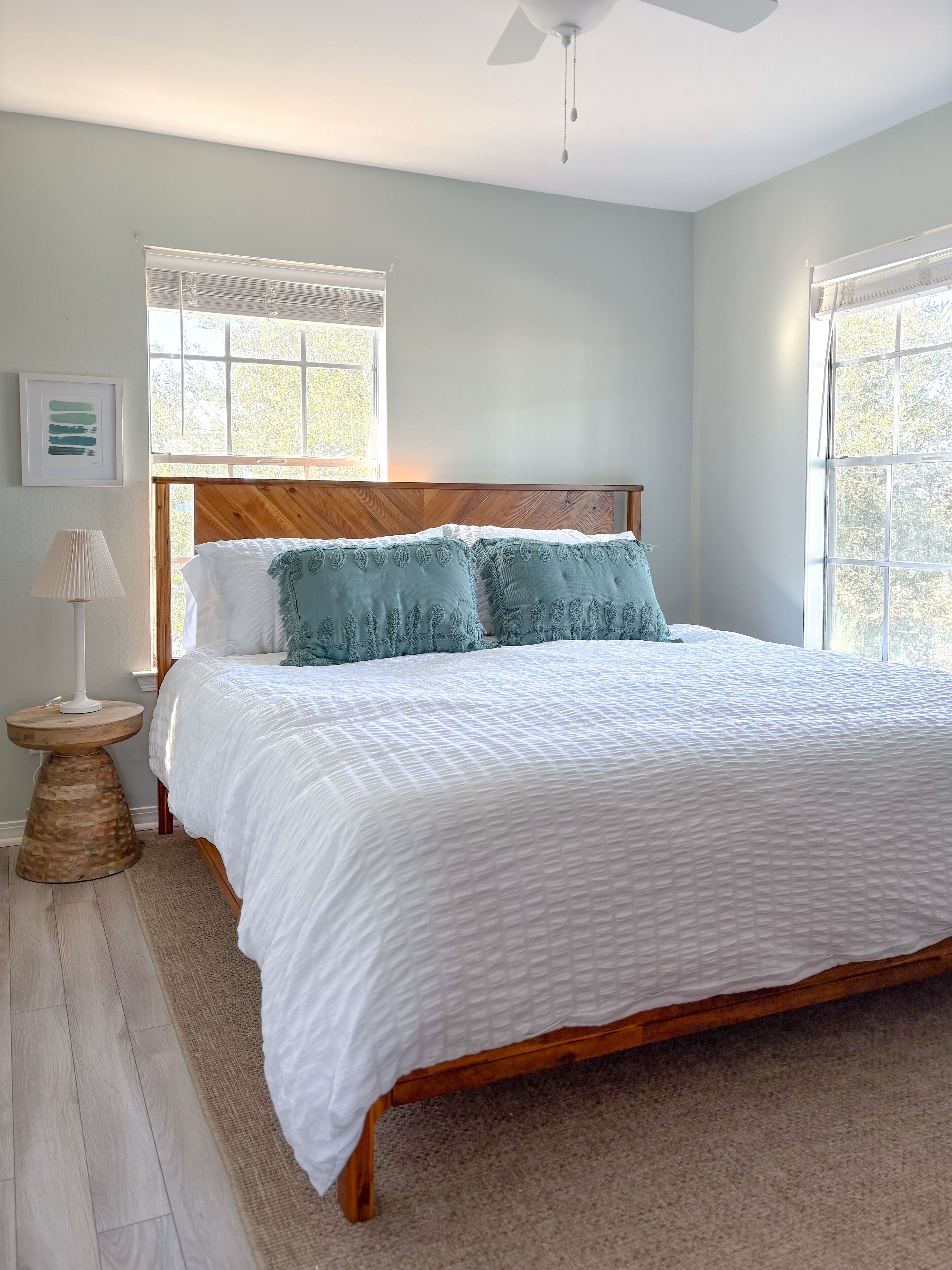 A bedroom with a wooden bed frame, white bedding, and two windows with blinds.