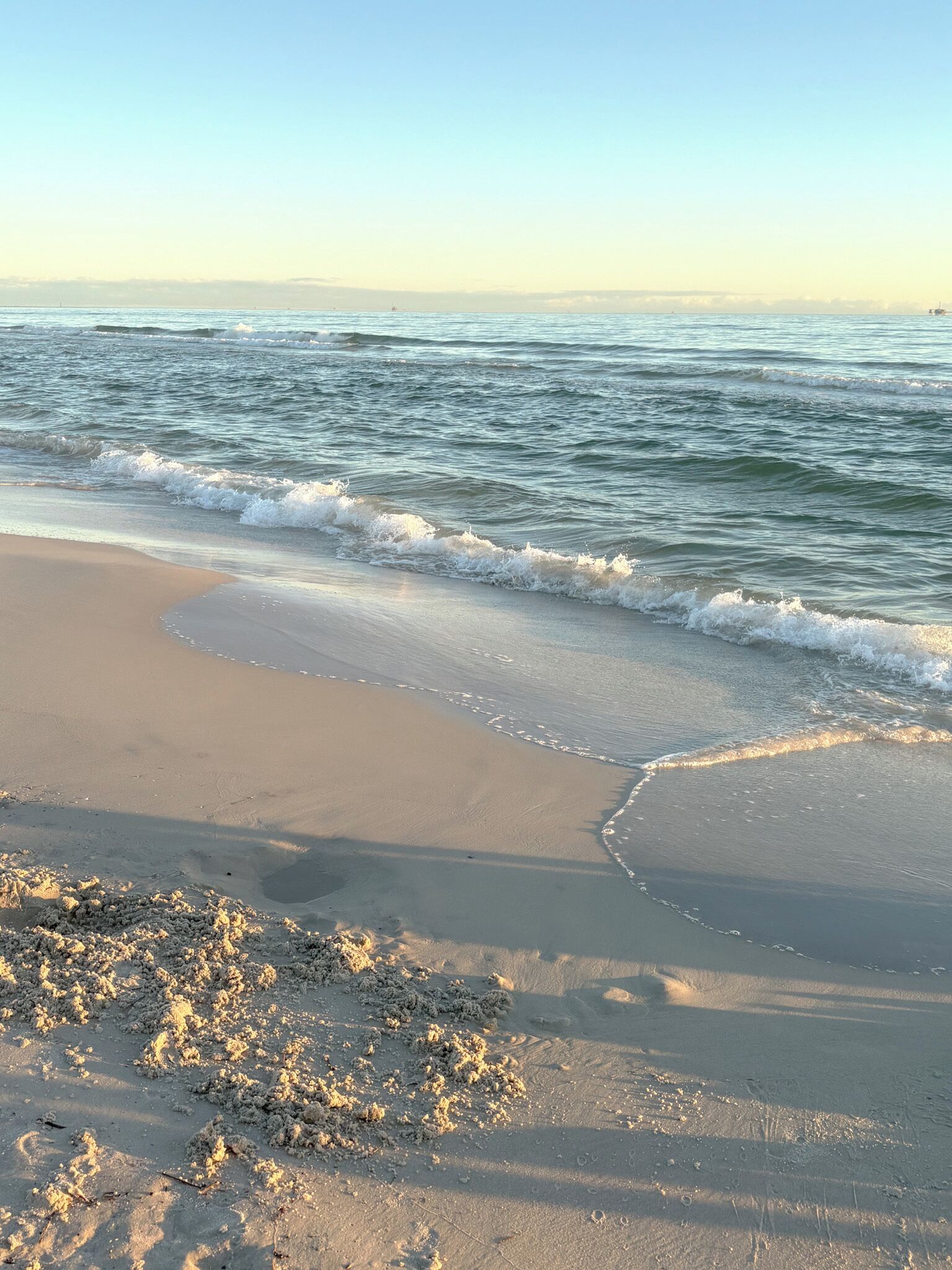 Een strand met golven die zachtjes op het strand komen en wegvloeien, heldere lucht en een rustige zee.