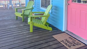 A porch with two green Adirondack chairs, a pink door, and a blue building in the background. - Walk to beach, restaurants, & shopping. Charming, pet-friendly 3BR bungalow.  (Dauphin Island)