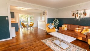 A spacious living room with a wooden floor, a brown leather sofa, a white coffee table, and a dining area with yellow chairs.