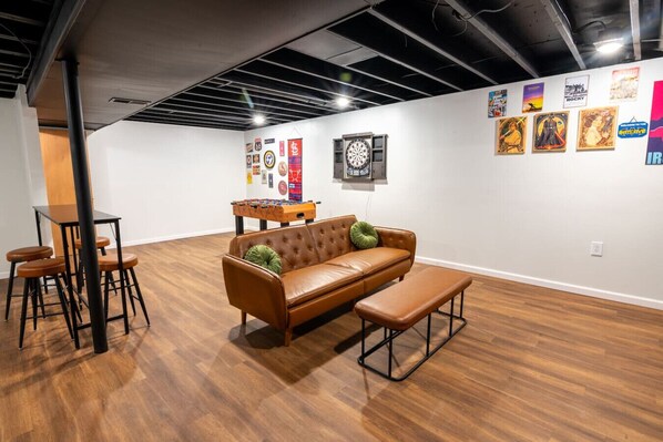 Une pièce avec un canapé en cuir marron, une table basse et une table de baby-foot. Il y a des affiches au mur et un jeu de fléchettes.