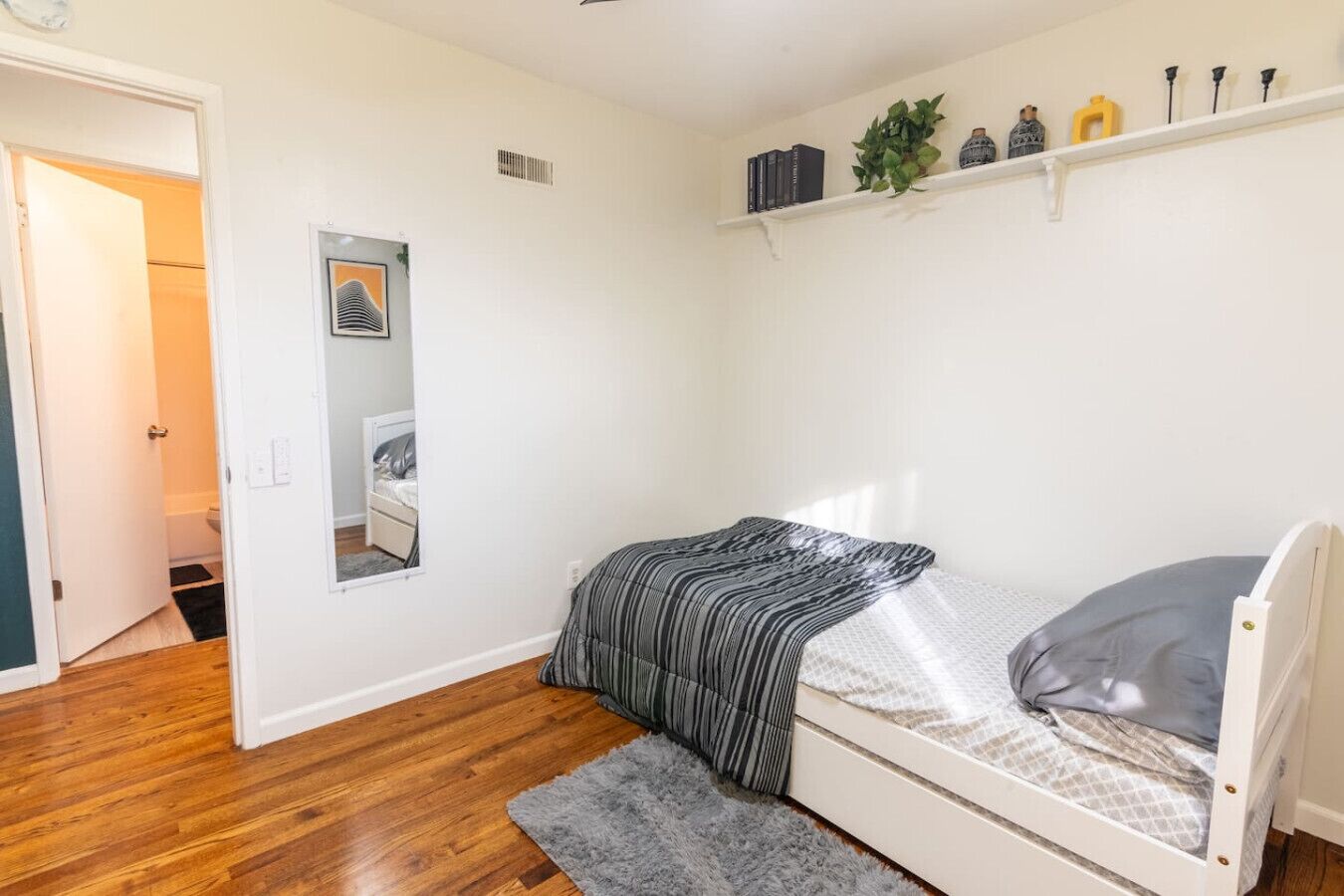 Un dormitorio con una cama, un espejo, una estantería con libros y objetos decorativos, y suelo de madera.