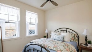 A bedroom with a bed, two bedside tables with lamps, a ceiling fan, and two windows with curtains. - The Red Mountain Suite @ HSRV (Ivins)