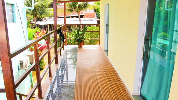 A corridor with a wooden railing, a potted plant, and a view of a building with a red roof. - Pousada Visual de Noronha (Fernando de Noronha)