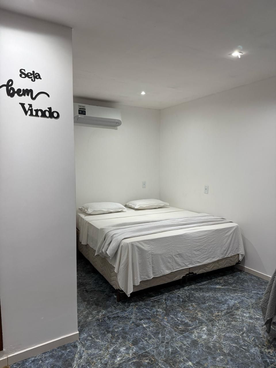 A bedroom with a bed, white walls, a ceiling light, and a marble floor.