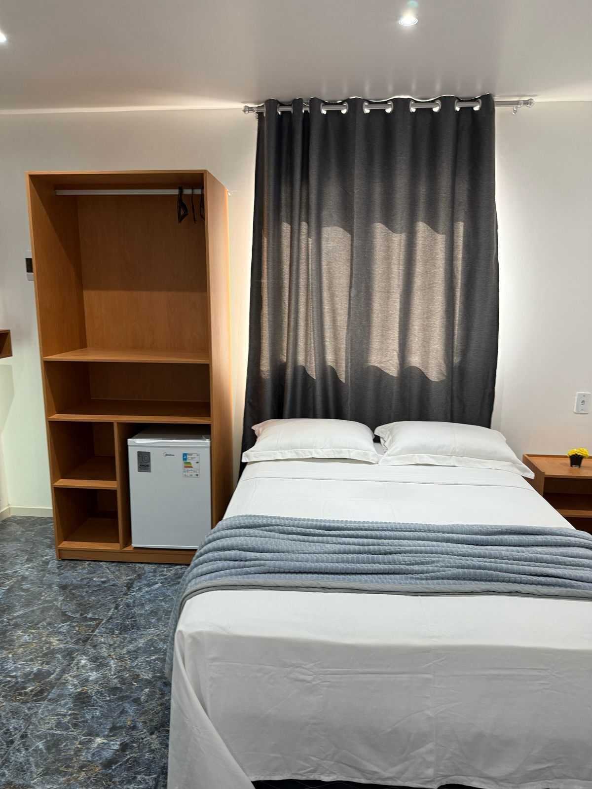 Une chambre à coucher comprenant un lit, une armoire, un petit réfrigérateur et une table de chevet avec une lampe.