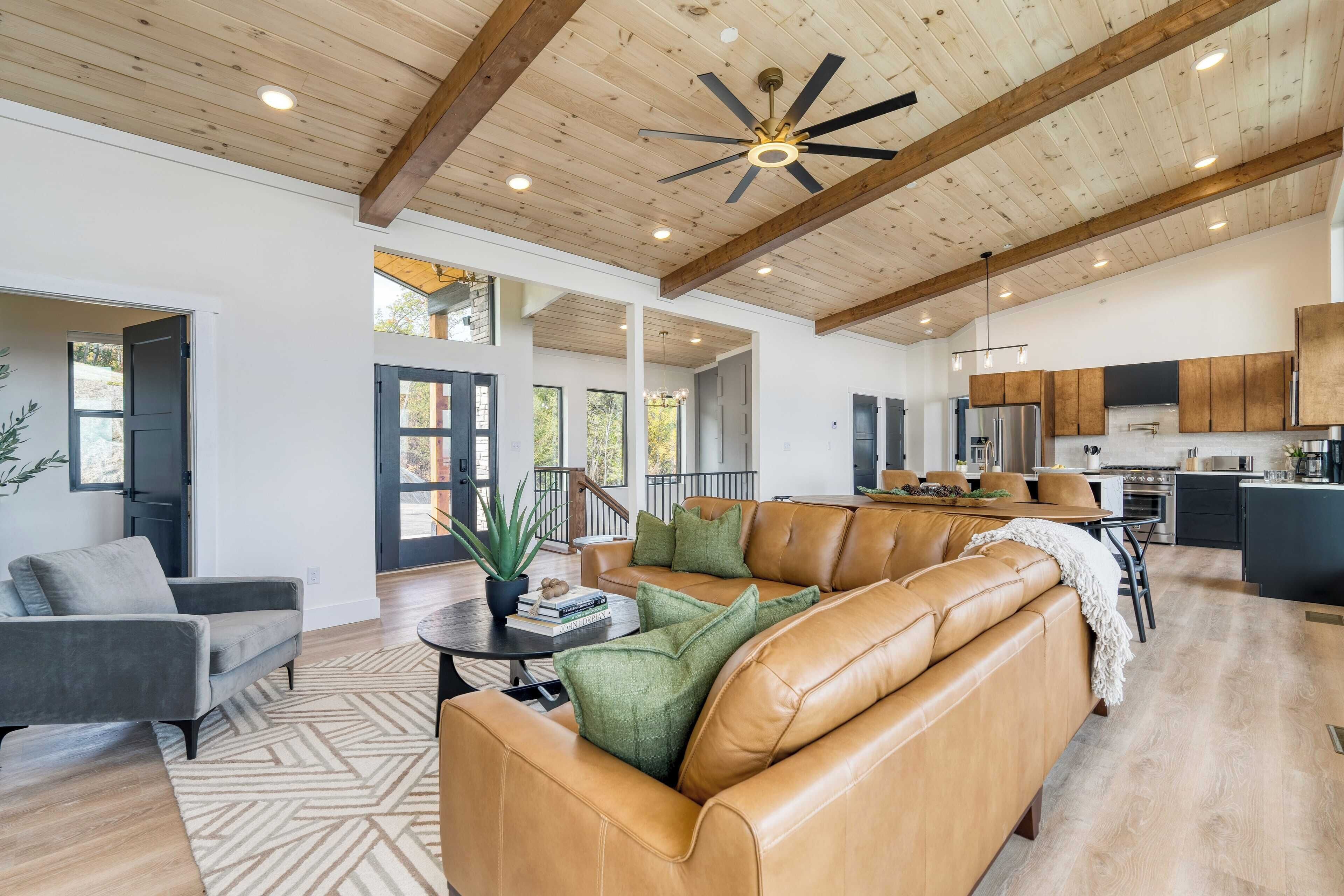 A spacious living area with a large leather sofa, a coffee table, and a potted plant. There is a kitchen in the background with modern appliances and a dining area.