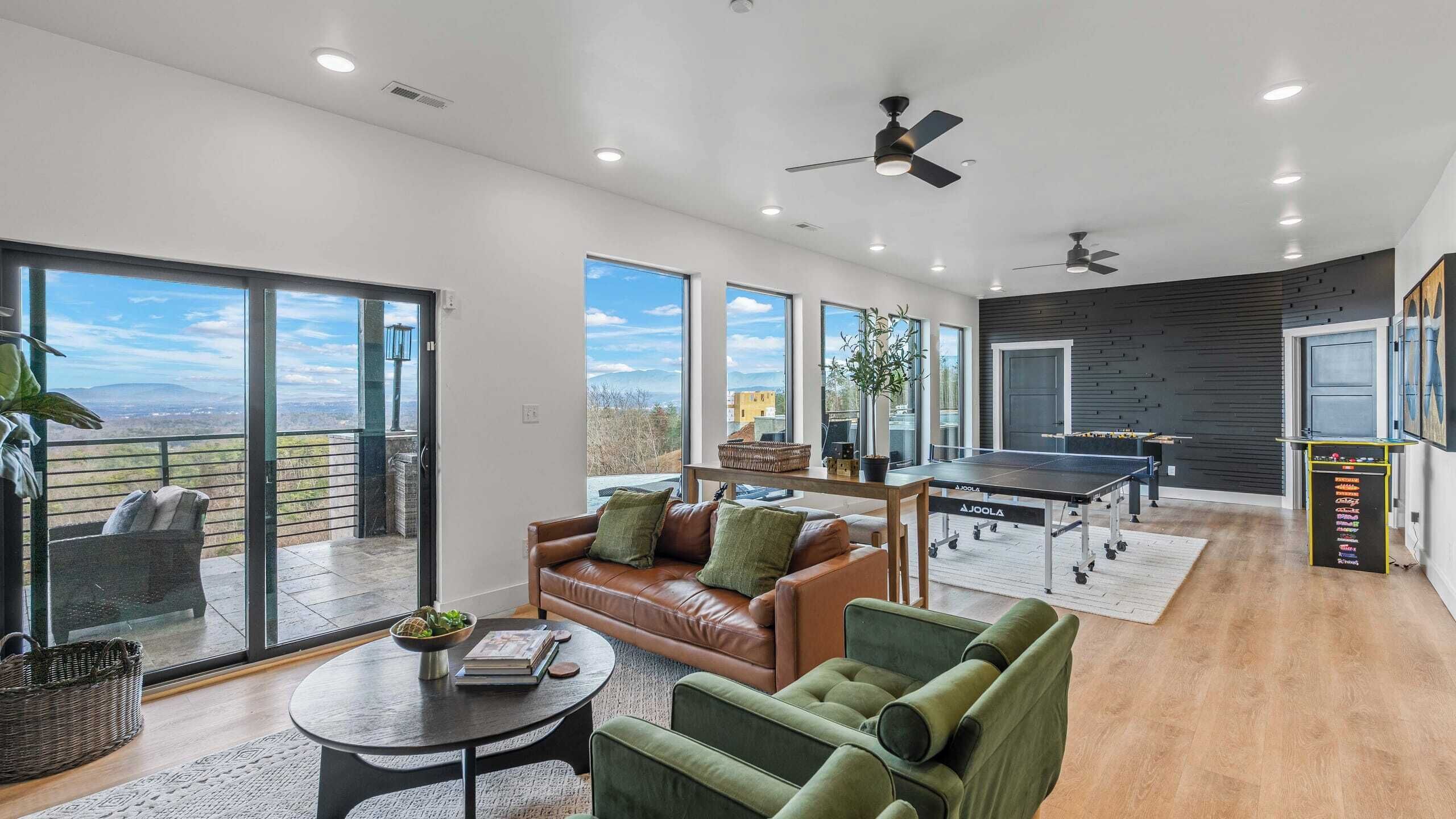 A spacious living area with a leather sofa, green armchairs, a ping pong table, and a large window offering a scenic view.