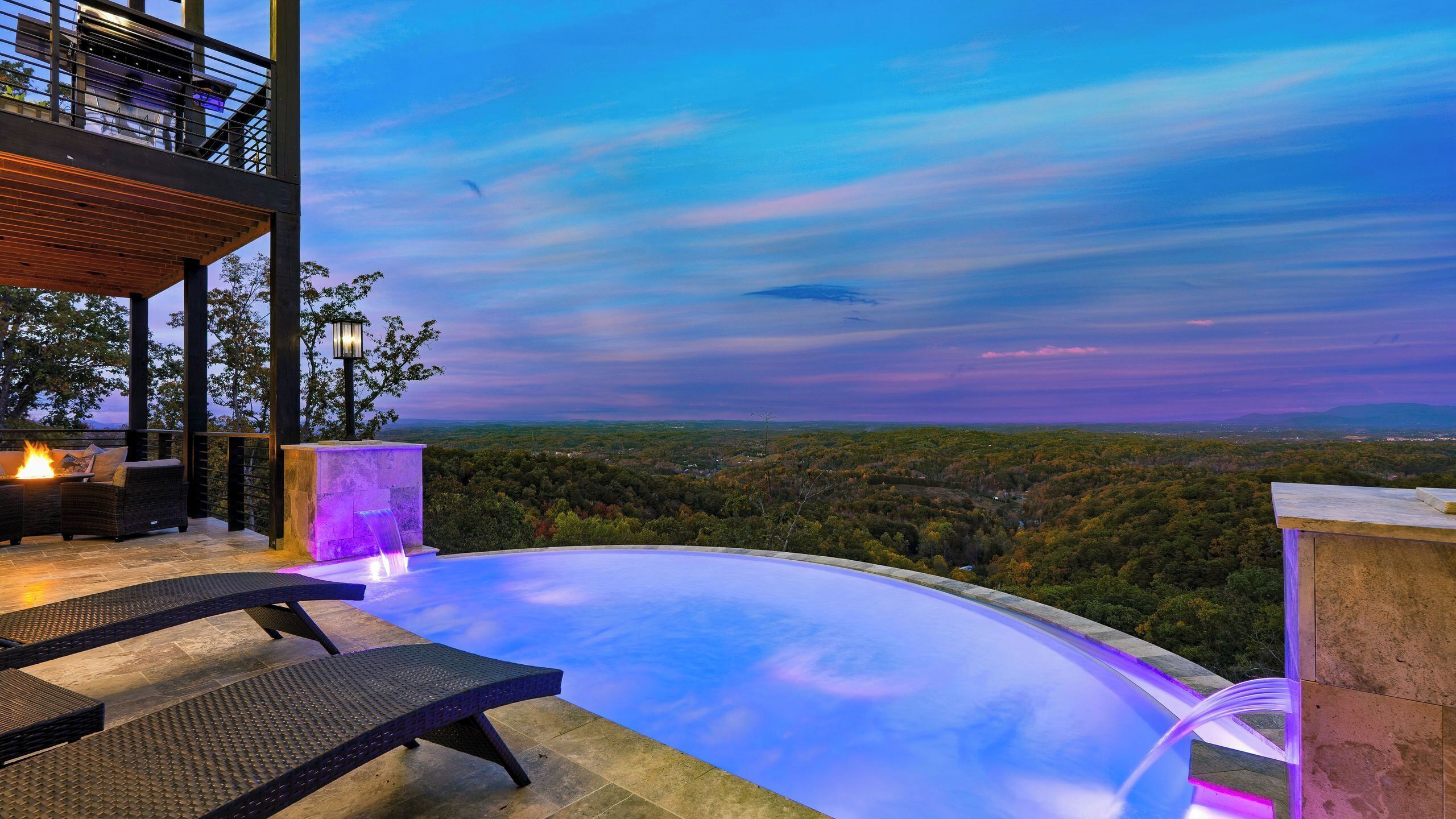 A modern outdoor infinity pool with a fire feature, lounge chairs, and a view of a forested landscape.