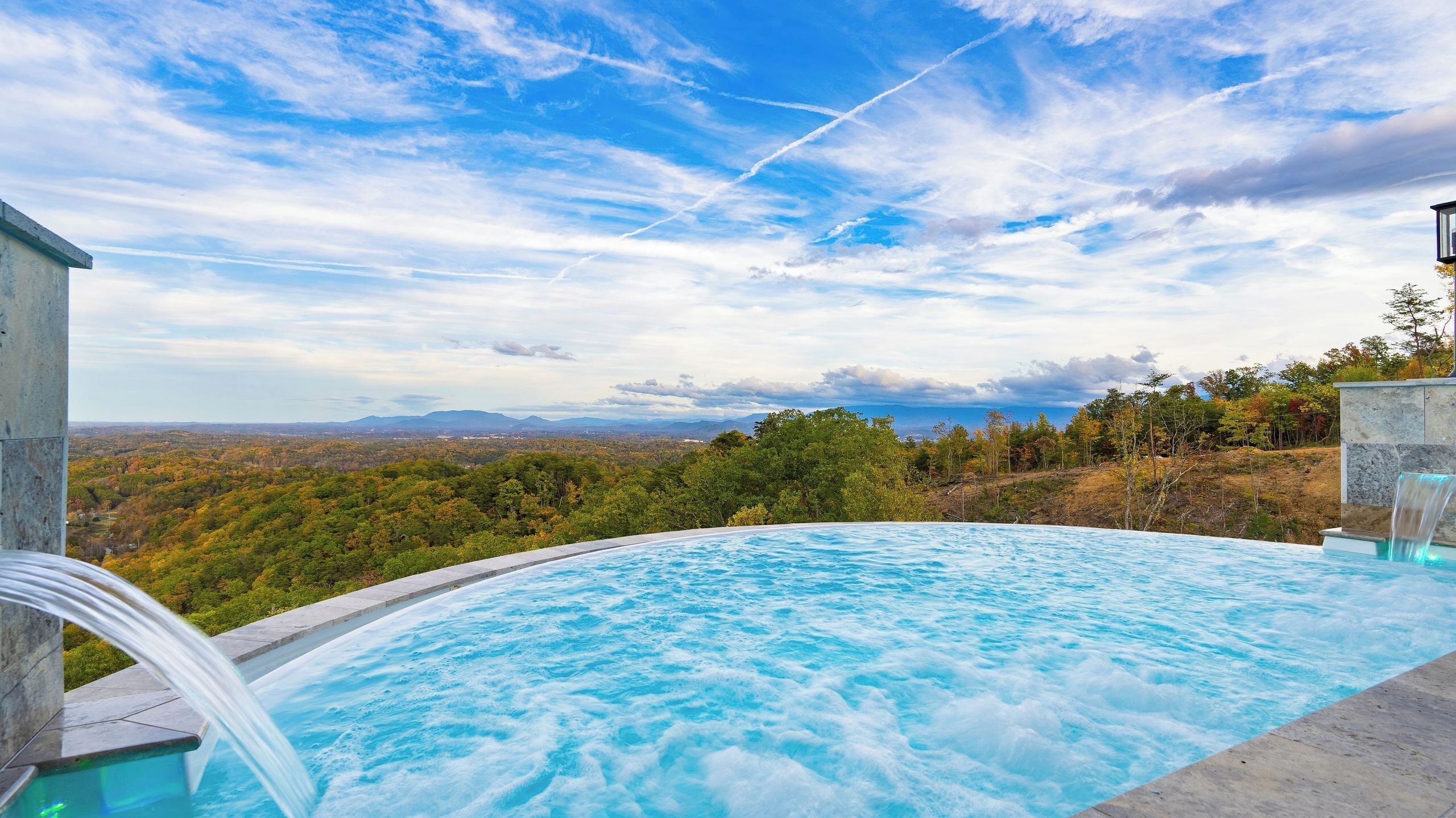 A hot tub with a waterfall feature, surrounded by a scenic view of a forested landscape and mountains.
