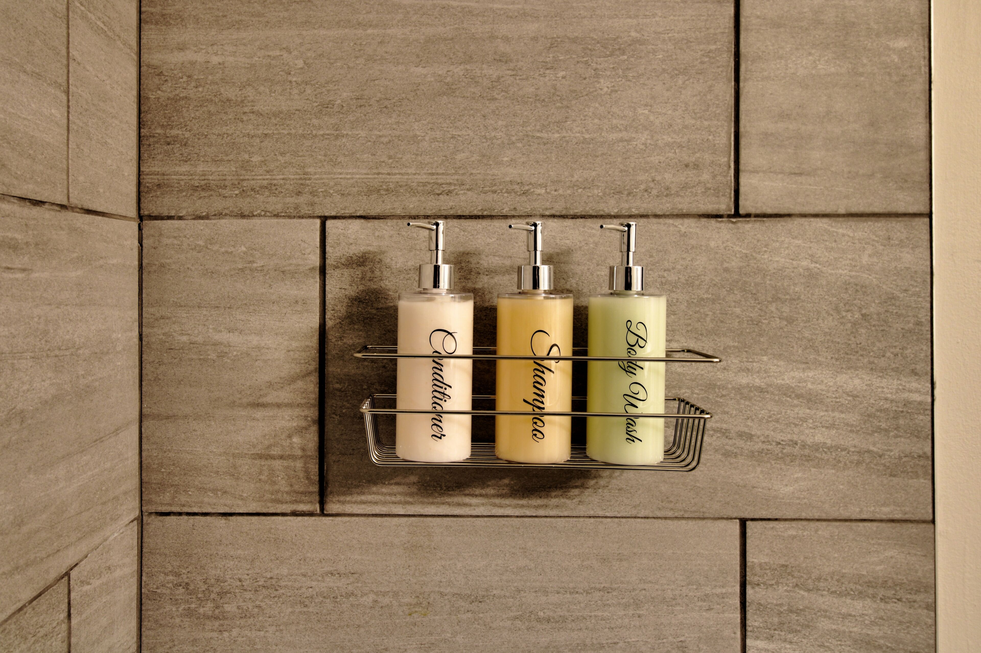 A bathroom with a tiled wall and a metal shelf holding three bottles of toiletries.