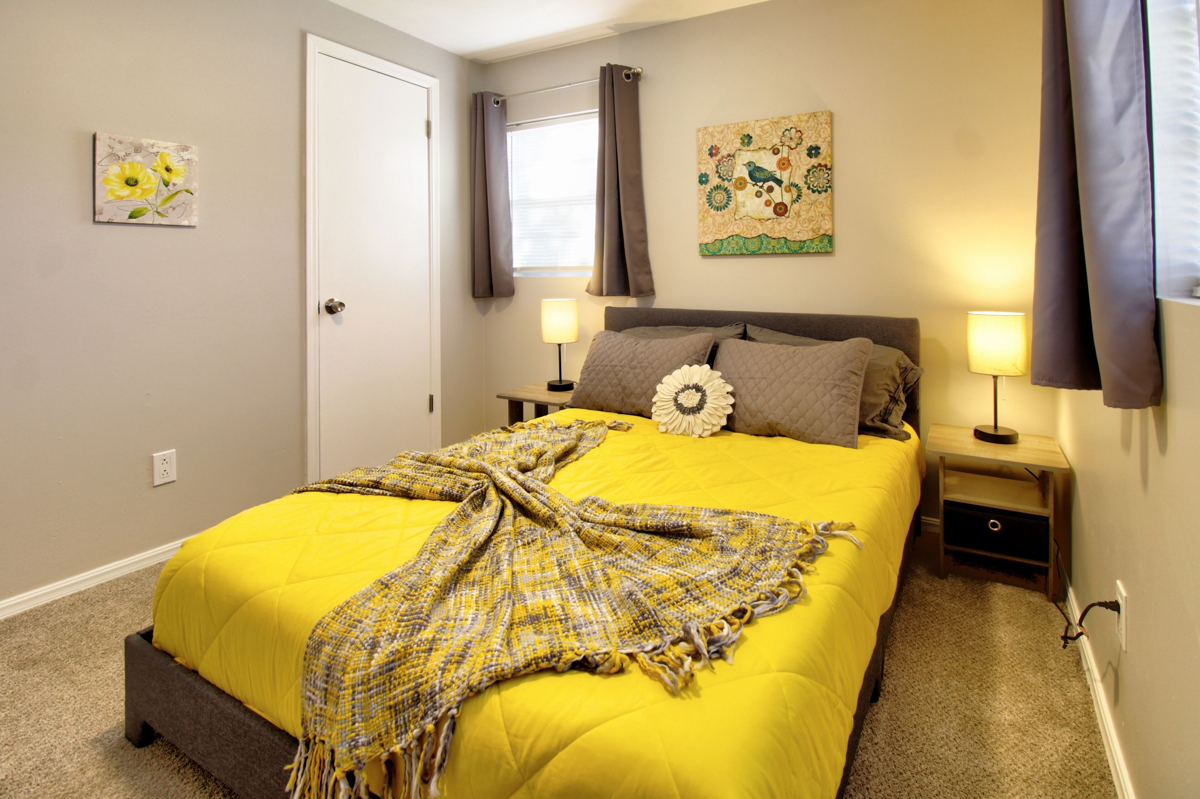Une chambre avec une couvre-lit jaune, une tête de lit grise, deux lampes de chevet et une œuvre d’art florale accrochée au mur.