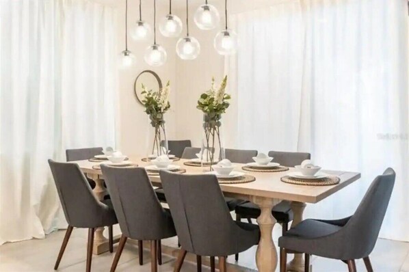 A dining area with a wooden table set for six, surrounded by six chairs. There are hanging pendant lights above the table, and two vases with flowers on either side.