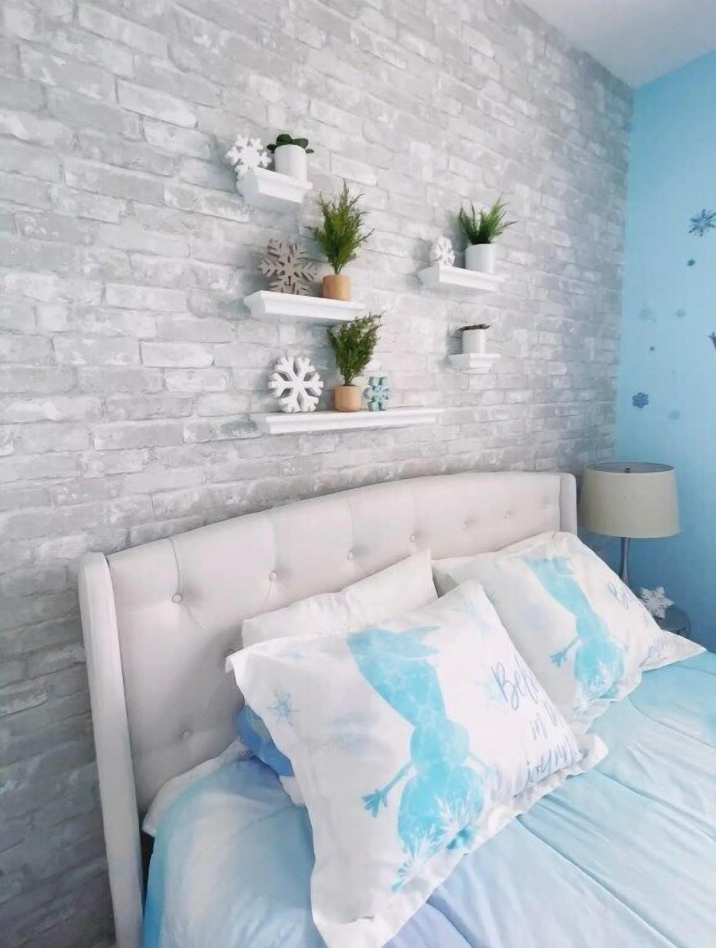A bedroom with a white brick wall, a tufted headboard, and a bed with blue bedding. There are white shelves with decorative items and plants.