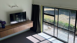 A room with a flat-screen TV on a wooden console, a large sliding glass door to a garden, and a small vase with flowers on the console. - ESCAPE TO THE COUNTRY (Kaitake)