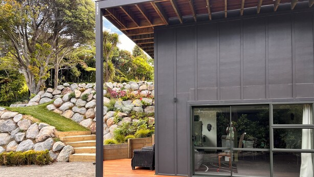 A modern house with a large glass door, a wooden deck, and a landscaped garden with rocks and plants. - ESCAPE TO THE COUNTRY (Kaitake)