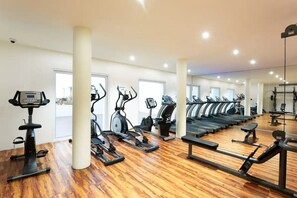 Ginásio com várias máquinas de cardio, soalho de madeira e grandes janelas.