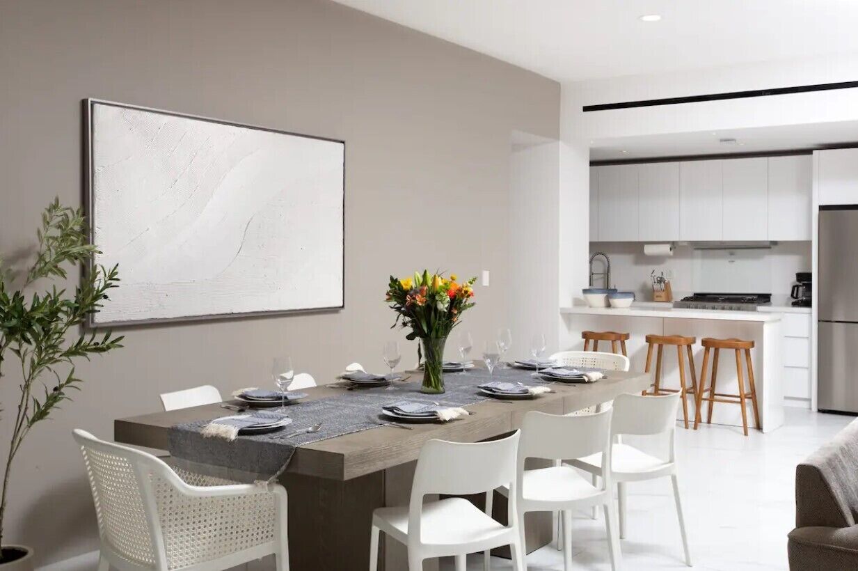 Un comedor moderno con una mesa larga dispuesta para seis comensales, sillas blancas, un jarrón de flores y un cuadro abstracto grande en la pared.