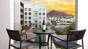 A balcony with two wicker chairs, a table, and a bottle of wine. The balcony overlooks a cityscape with buildings and a mountain in the background.