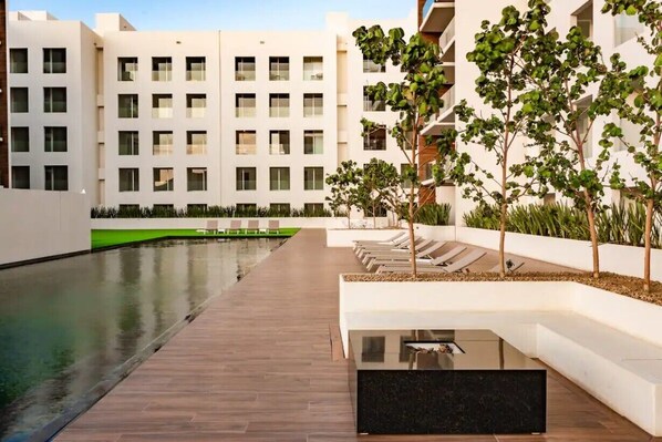 Um edifício moderno com vários andares, piscina e área ajardinada com árvores e assentos.