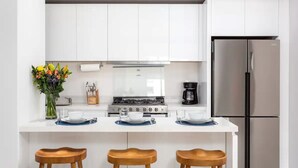 A modern kitchen with white cabinets, a stainless steel refrigerator, a gas stove, and wooden stools.