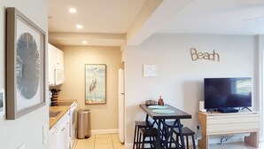 Un espace de vie compact comprenant un coin cuisine, un coin repas avec une table et des chaises, un téléviseur posé sur un meuble et une œuvre d’art accrochée au mur.