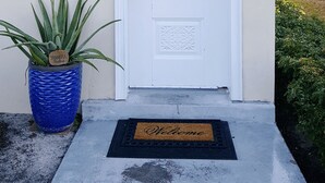 Une porte blanche avec une plaque portant le numéro « 582 », un pot de fleurs orné d’une étiquette indiquant « MERCI », et un paillasson avec l’inscription « BIENVENUE ».