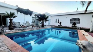 A swimming pool with lounge chairs and umbrellas, surrounded by a white building with large windows and a stone pathway. - Villa Dar sukar  (Marrakech)