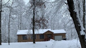 Une maison à deux étages, dont le toit est recouvert de neige, avec un garage, le tout entouré d’une forêt enneigée.