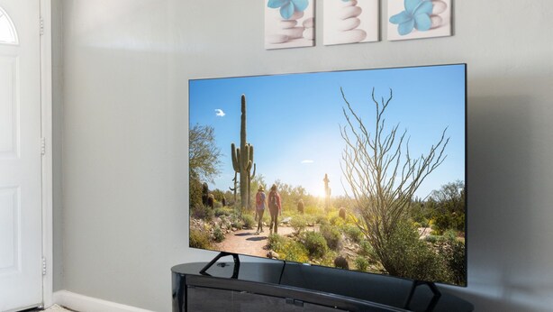 Televisão na parede exibe paisagem desértica com cactos e duas pessoas caminhando.