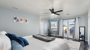 Quarto com cama, ventoinha no teto, escrivaninha e vista através de uma porta de vidro.