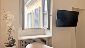 A room with a white chair, a potted orchid, a window with shutters, and a flat-screen TV mounted on the wall. - Centro Corso Venezia - Chic & Quiet Apartment in M (Milan)