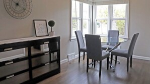 A dining area with a glass table surrounded by chairs, a black console table with drawers, a wall-mounted decorative piece, and a window with white blinds.