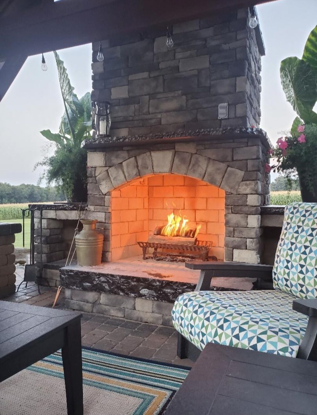 An outdoor stone fireplace with a fire, surrounded by outdoor furniture and a view of a field.