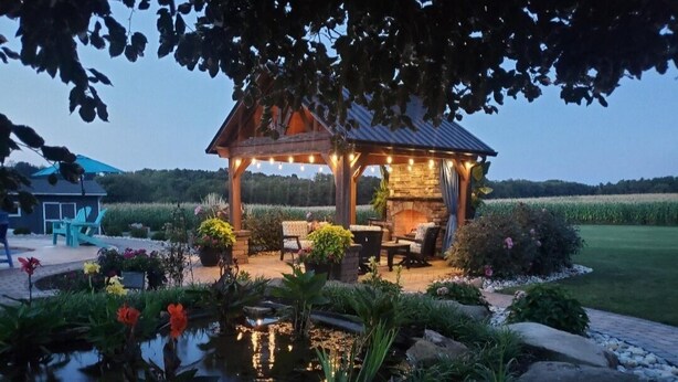 Ein Gazebo mit Kamin aus Stein, umgeben von einem Garten mit Teich und unterschiedlichen Pflanzen.