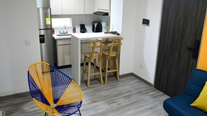A compact kitchen with a refrigerator, stove, and sink. There are wooden chairs and a yellow chair. A blue sofa with a yellow pillow is in the living area. - Spectacular Apartment in the Zona Rosa - Stratum 5 (Manizales)