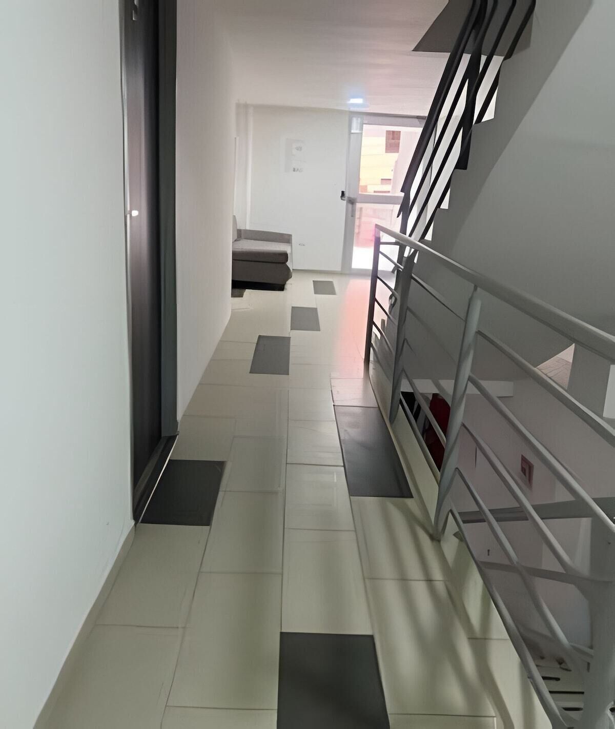 Un pasillo con una escalera a la derecha que conduce a un piso superior. El suelo presenta un diseño de baldosas claras y oscuras.