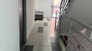 Un pasillo con una escalera a la derecha que conduce a un piso superior. El suelo presenta un diseño de baldosas claras y oscuras.