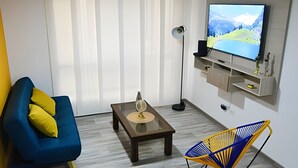 A living room with a blue sofa, a coffee table, a floor lamp, a TV mounted on the wall, and a chair with yellow and blue accents. - Spectacular Apartment in the Zona Rosa - Stratum 5 (Manizales)
