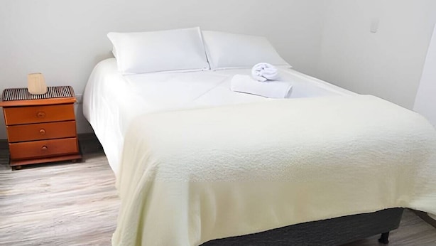 Un lit bien fait, avec du linge de lit blanc et une serviette pliée, une table de chevet en bois avec une lampe, et un matelas de couleur foncée.