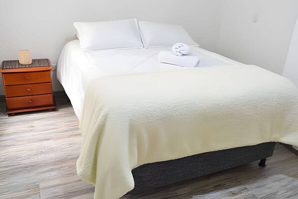 A neatly made bed with white linens and a folded towel, a wooden nightstand with a lamp, and a dark-colored mattress.