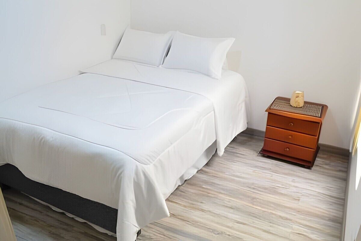 Una cama bien tendida con ropa blanca en una habitación con piso de madera y una mesita de noche de madera con cajones.