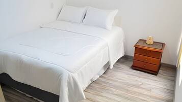 Una cama bien tendida con ropa blanca en una habitación con piso de madera y una mesita de noche de madera con cajones.