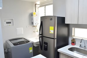 A compact kitchen with a stainless steel refrigerator, a built-in oven, a sink, and a window with blinds.