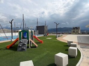 A playground with slides and climbing structures, surrounded by artificial grass and concrete seating.